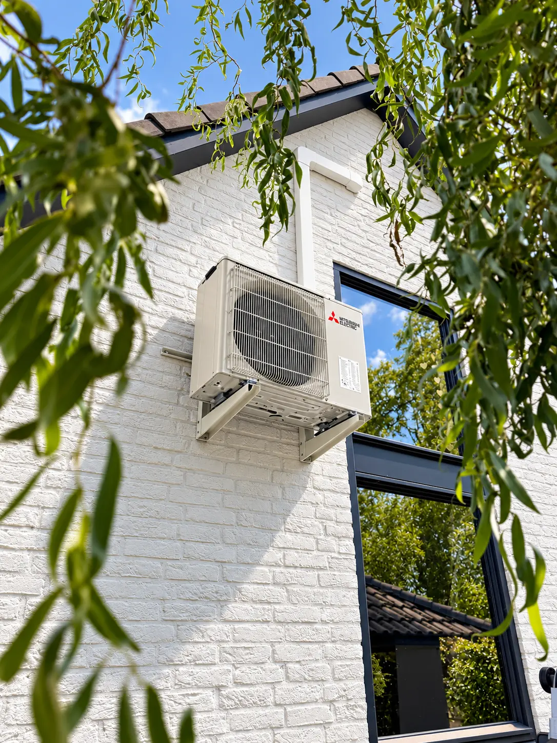 Mitsubishi Electric buitenunit op consoles hoog aan een witte gevel tussen het groen